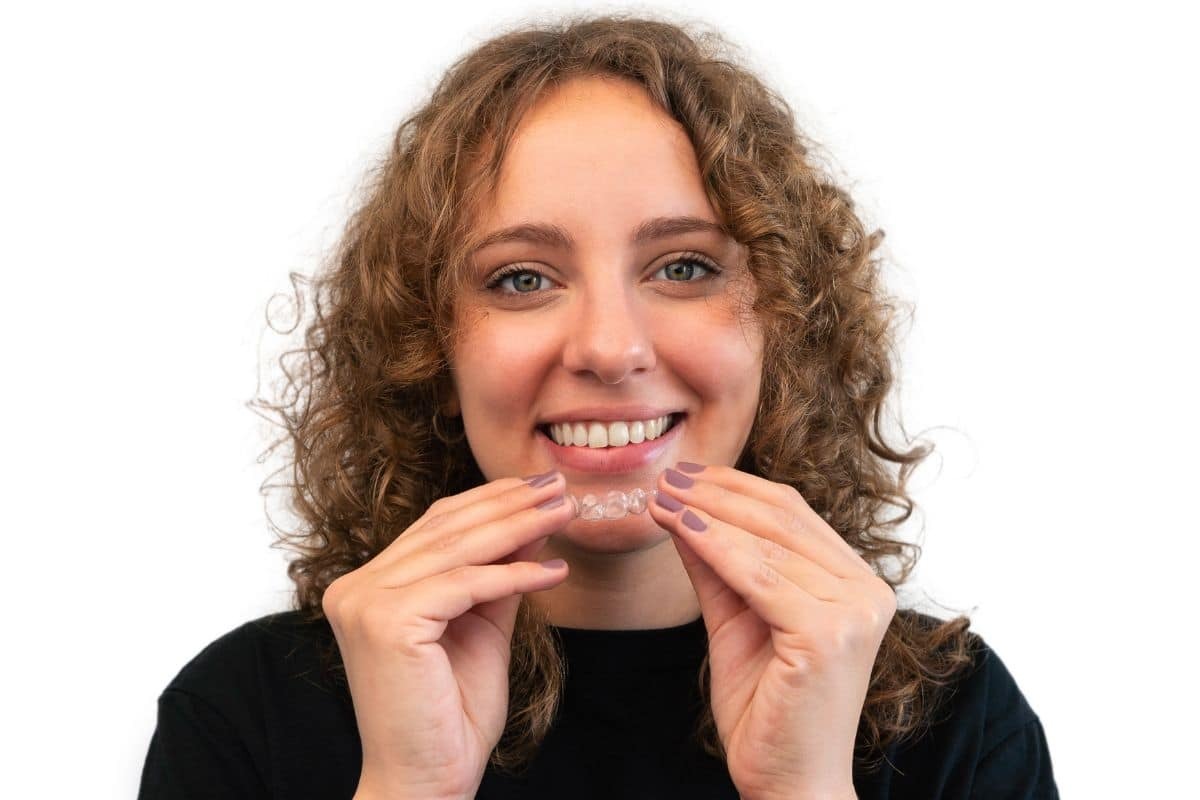 Young woman holding clear Invisalign aligner tray and smiling at camera.